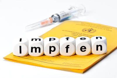 Bild vergrößern: Spritze mit Impfstoff auf Internationalen Impfpass. Impfen geschrieben mit Würfeln.