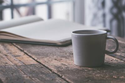 Bild vergrern: Ein rauer Holztisch, darauf steht vorne eine mittlere graue Keramik Tasse mit Kaffee im Hintergrund ein aufgeschlagenes Buch mit weien leeren Seiten.